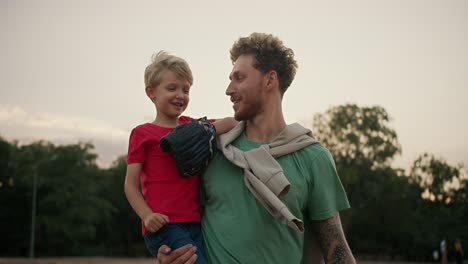un niño rubio feliz con una camiseta roja sostiene una pelota de béisbol en sus manos y la lanza al guante de su padre con una camisa verde. retrato de un niño y su padre jugando béisbal en el parque