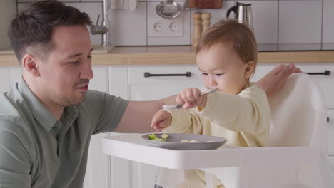 Linda-Niña-Sentada-En-Una-Silla-Alta,-Sosteniendo-Un-Tenedor-Y-Jugando-Con-La-Comida-Mientras-Su-Padre-La-Miraba