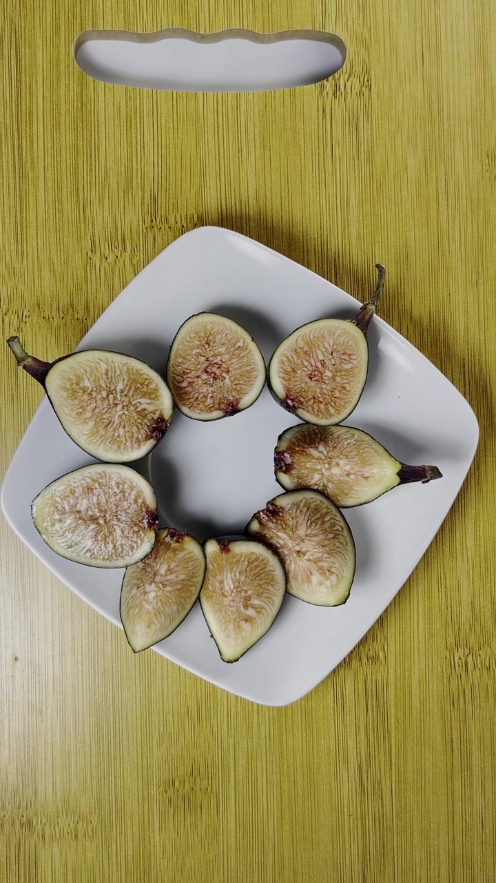 Fresh figs are sliced and arranged on a white plate over a wooden surface. Pale flesh green skin and detailed seeds create a simple food composition viewed from above.
