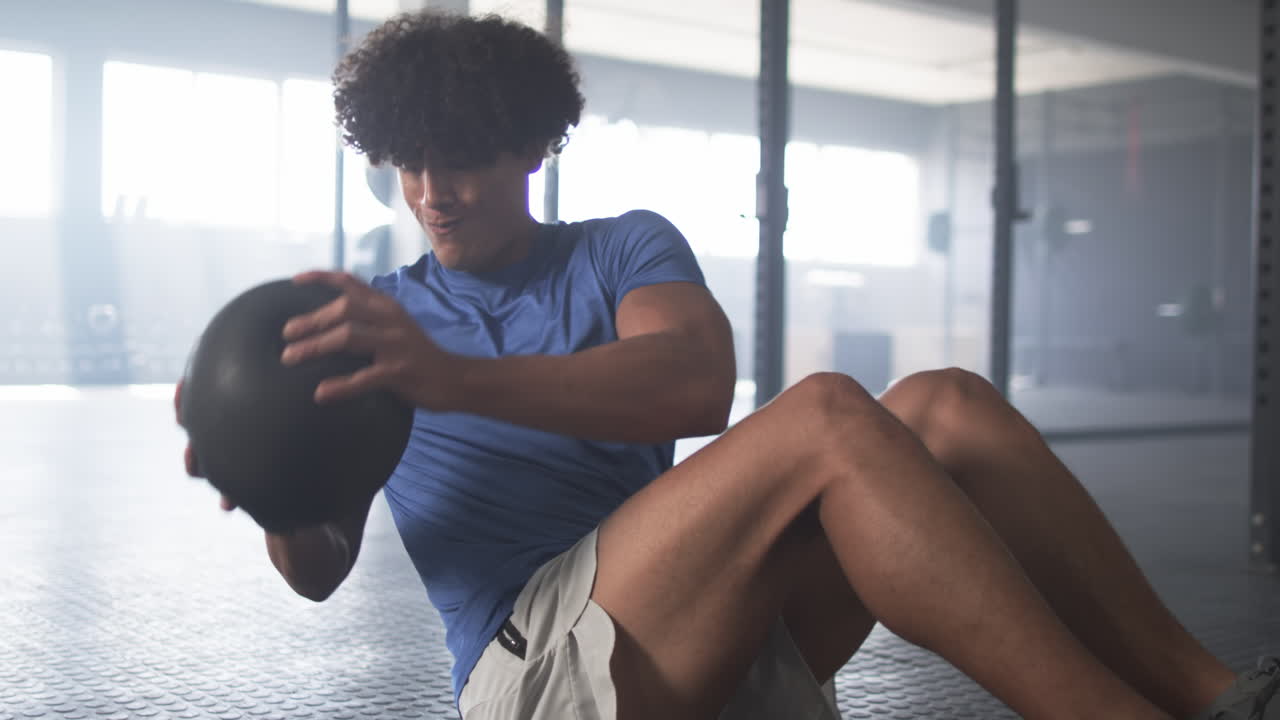 Exercising in gym, man doing sit-ups with medicine ball, focusing on fitness