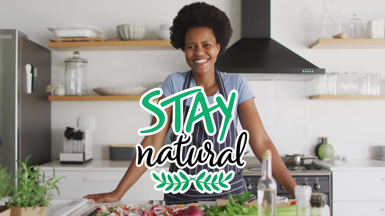 Animation of stay natural text over happy biracial woman with apron at home