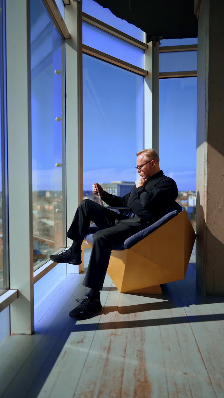 Businessman sits in the chair in the comfortable office near the panoramic window. Man works on laptop and enjoys the city view. Vertical video.