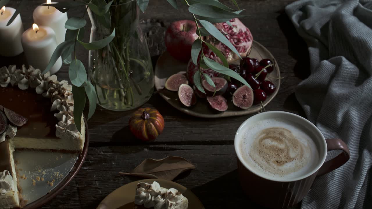 Rustic still life features flowers in vase, burning candles, figs, cherries, pomegranate, pumpkin, cake, coffee, and cozy textile on wooden table, autumn mood