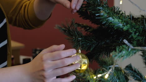 Woman-hands-placing-yellow-ornament-setting-up-christmas-tree-with-lights-tight-shot