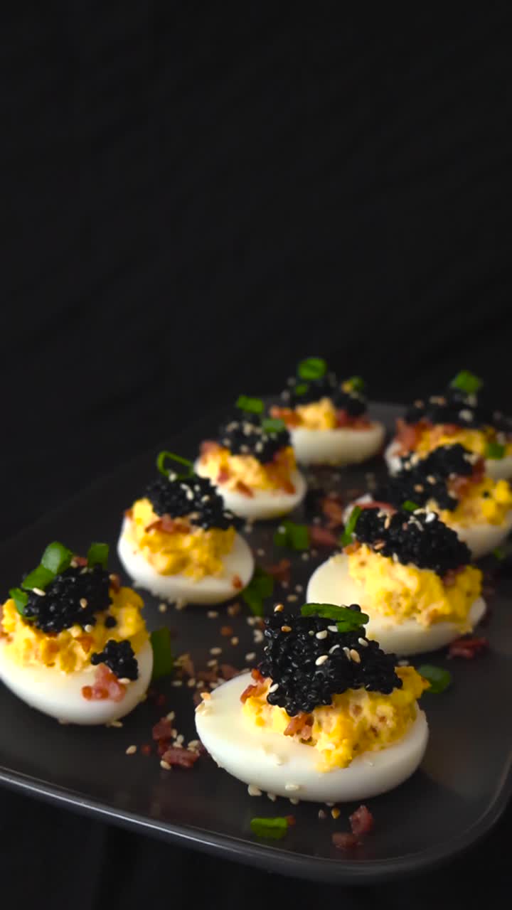 Lækker gourmetforret af fyldte æg med sort kaviar, baconstykker og purløg elegant præsenteret på en mørk tallerken, der roterer blidt mod en sofistikeret sort baggrund.