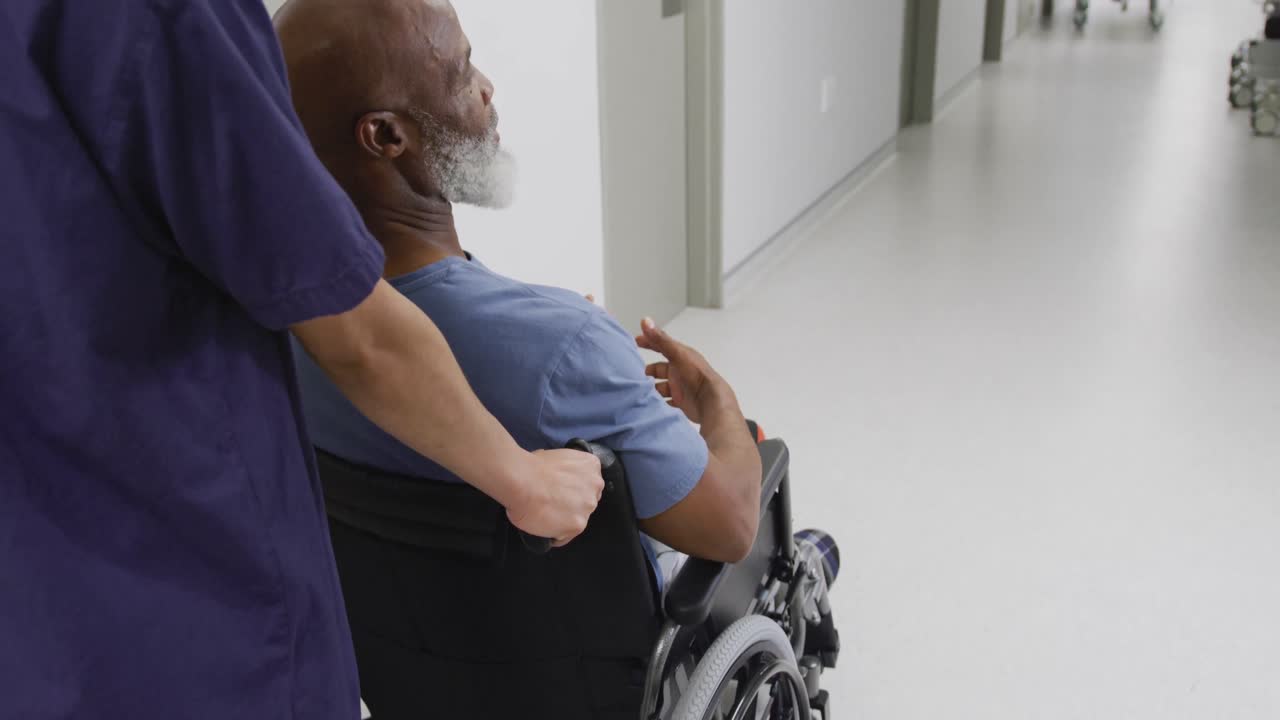 Diverse female doctor pushing senior male patient in wheelchair down hospital corridor