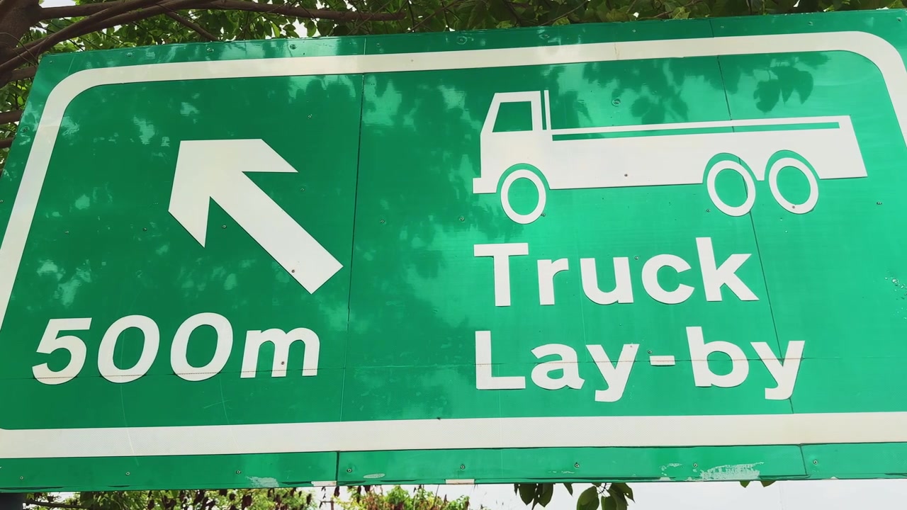 Langsame Kamerafahrt eines Lastwagens an einem Wegweiser, der einen Parkplatz voraus entlang einer Autobahn-Verkehrsachse anzeigt.