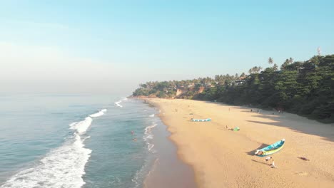 golden tropical sandy beach with moored fishing boats and tourists out walking in varkala, kerala, india - aerial low angle fly-over