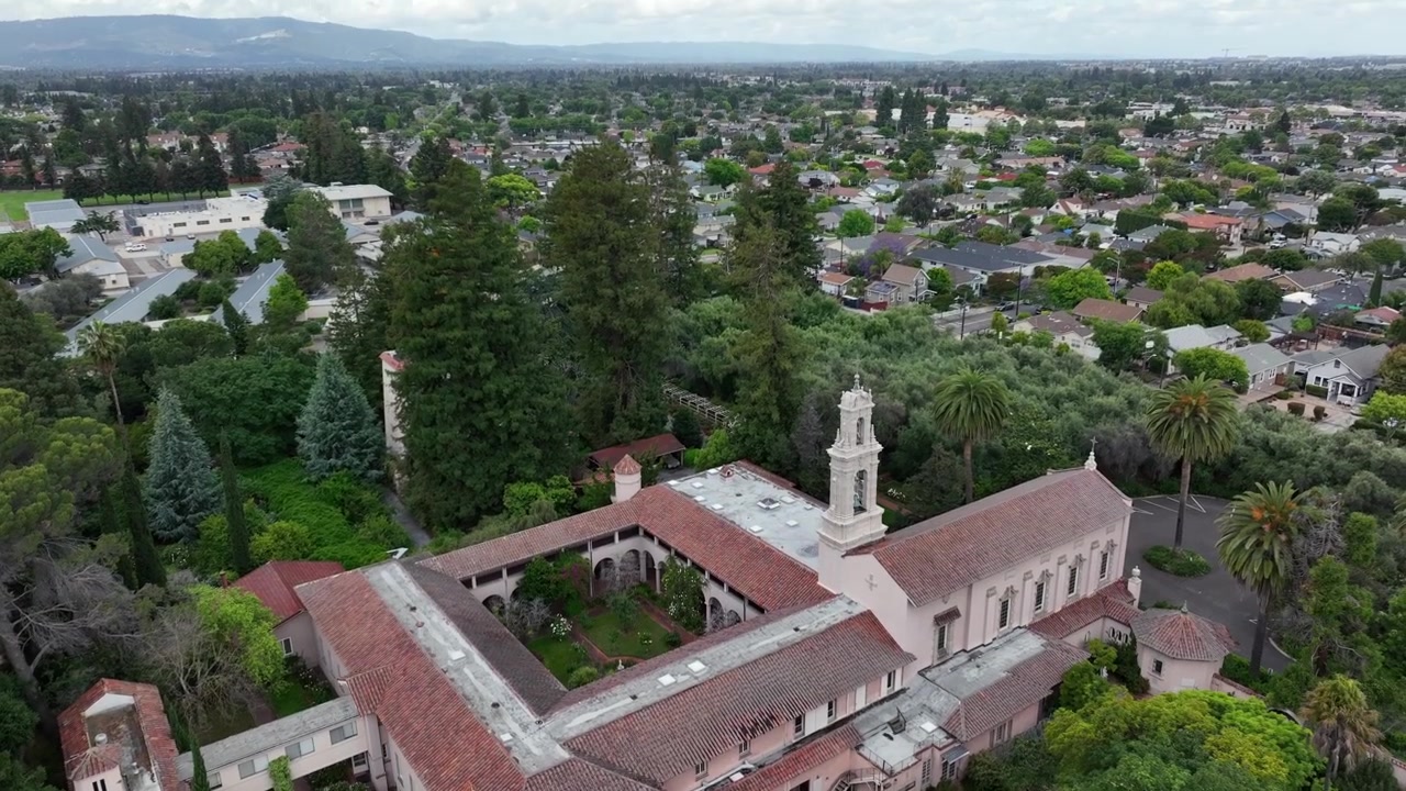 Iglesia de misión española con campanario blanco, tejados de tejas rojas y patio, anidada entre imponentes coníferas junto a calles residenciales en Santa Clara, California, EE. UU. Toma aérea de dron con inclinación ascendente