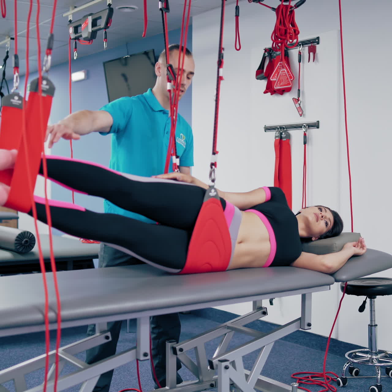 Young trainer explains the client how to do workout after the injury in rehab center. Woman is lying on special table and doing recovery exercises with suspension ropes with the help of instructor indoors.