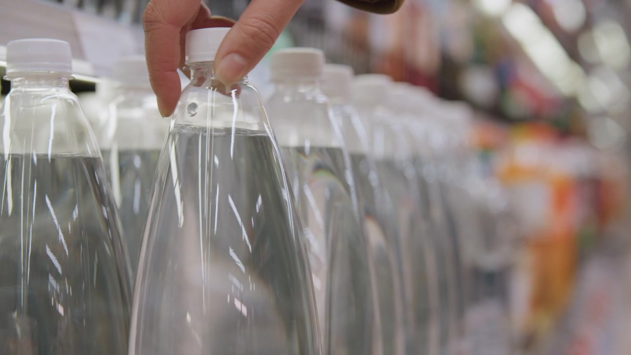 Close-up van rijen doorzichtige waterflessen op een schap, glimmende doppen en reflecterende oppervlakken, hand van een shopper reikt naar een gekoelde fles, heldere supermarktverlichting en wazige kleurrijke gangpadachtergrond.