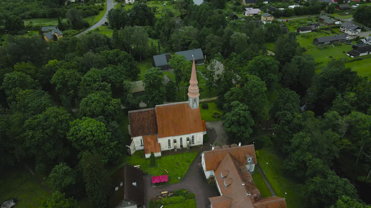 Aerial drone orbit view of old medieval church in Palamuse small village. Timeless stone architecture, red rooftops nestled in lush countryside greenery, peaceful landscape during overcast summer day.