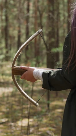 purple haired teenage girl holds crossbow walking across green wood backside view slow motion. theme play with medieval items. leisure and activity