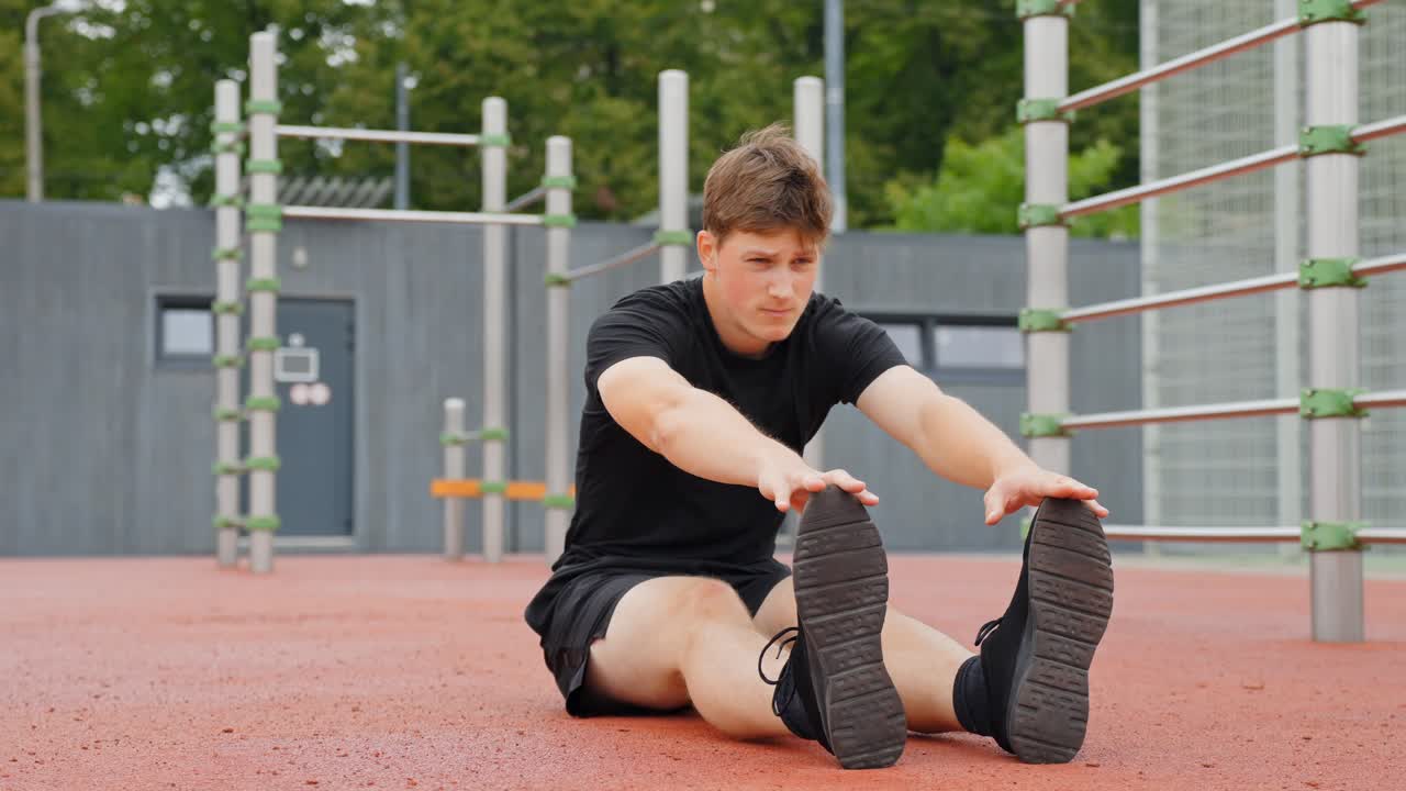 Fixed shot of a man sitting on the ground in an outdoor sports area, stretching his legs with arms extended