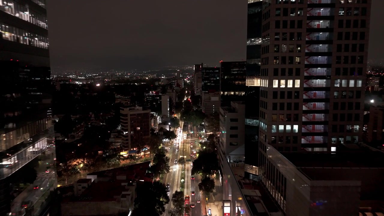Vuelo nocturno aéreo sobre una avenida arbolada y torres de la Ciudad de México