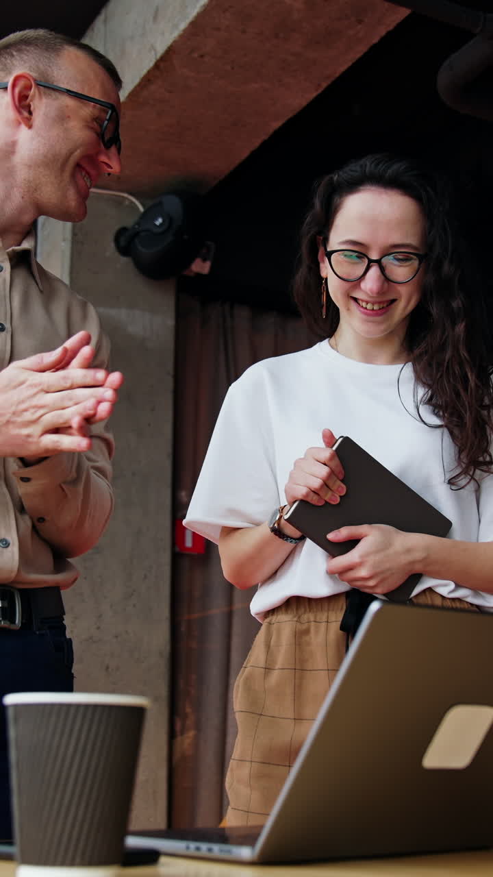 Cheerful office workers in the spacious office. Colleagues stand near the desk with laptop and discuss project. Low angle view. Vertical video