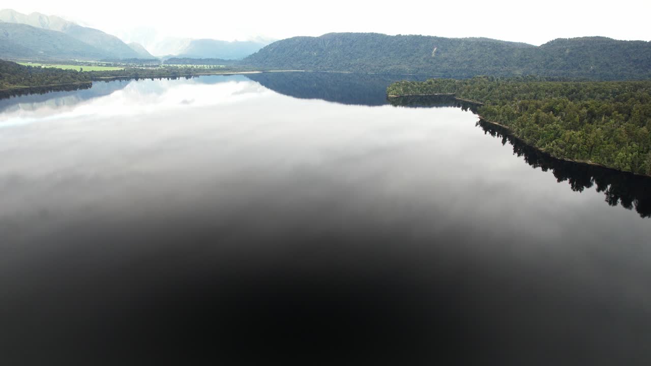 Specular Waters Of Lake Mapourika Surrounded By Lush Forest And Distant Hills In Fog. West Coast Of New Zealand's South Island. wide aerial shot