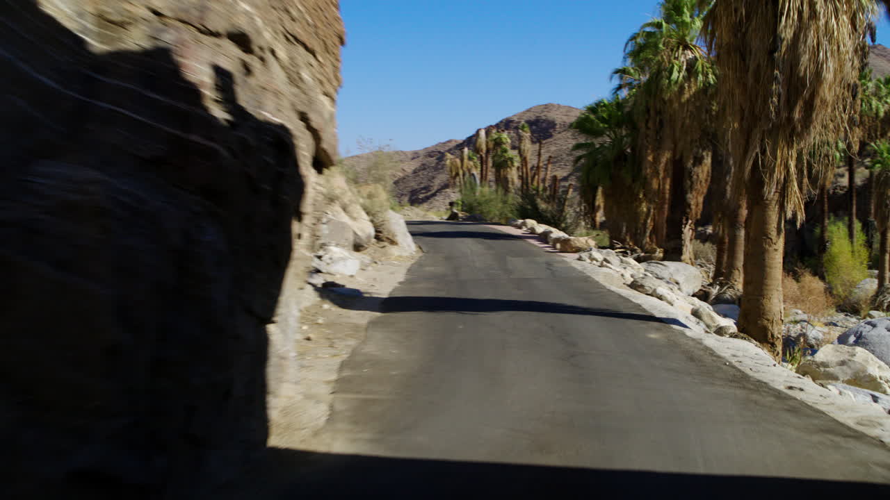 POV driving through “The Rock Drive-thru” at Indian Canyon in Palm Springs, California, surrounded by towering rock walls and palm trees, under bright sunshine and a clear blue sky