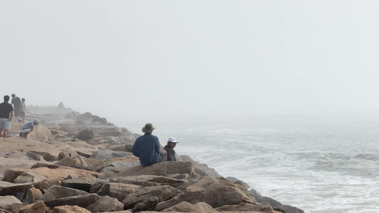 People Watching Waves Crash on Rocky Shore in Foggy Conditions