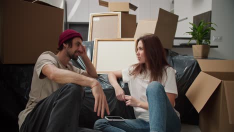 un hombre moreno con paja en un sombrero rojo y una camiseta beige está hablando con su novia morena en una camisetta blanca sentado en el suelo apoyado en el sofá en una cubierta de plástico negro y entre un gran número de cajas en un nuevo apartamento después de mudarse