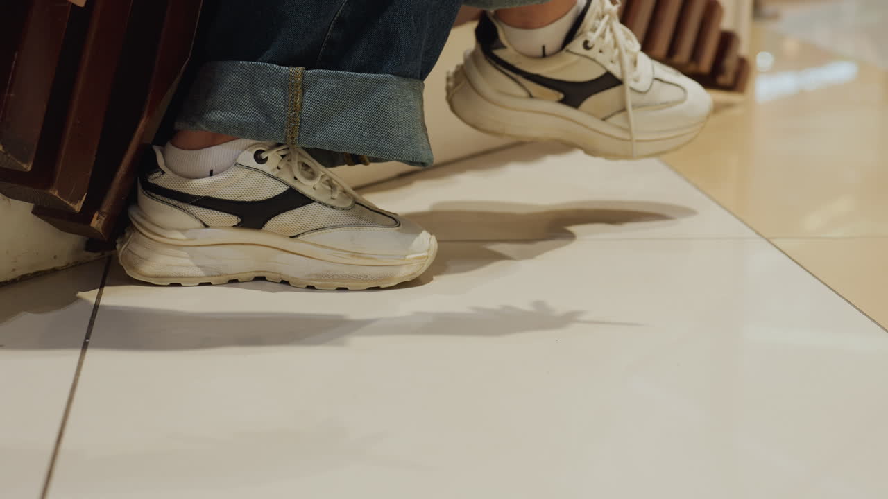 타일 위로 스니커즈가 클로즈업되어 발을 내딛고, 떨어진 물건을 조심스럽게 줍기 위해 손을 뻗는 모습이에요. 그림자와 윤기 있는 바닥에 은은하게 비치는 반영, 캐주얼한 복장과 느리고 신중한 움직임이 돋보여요.