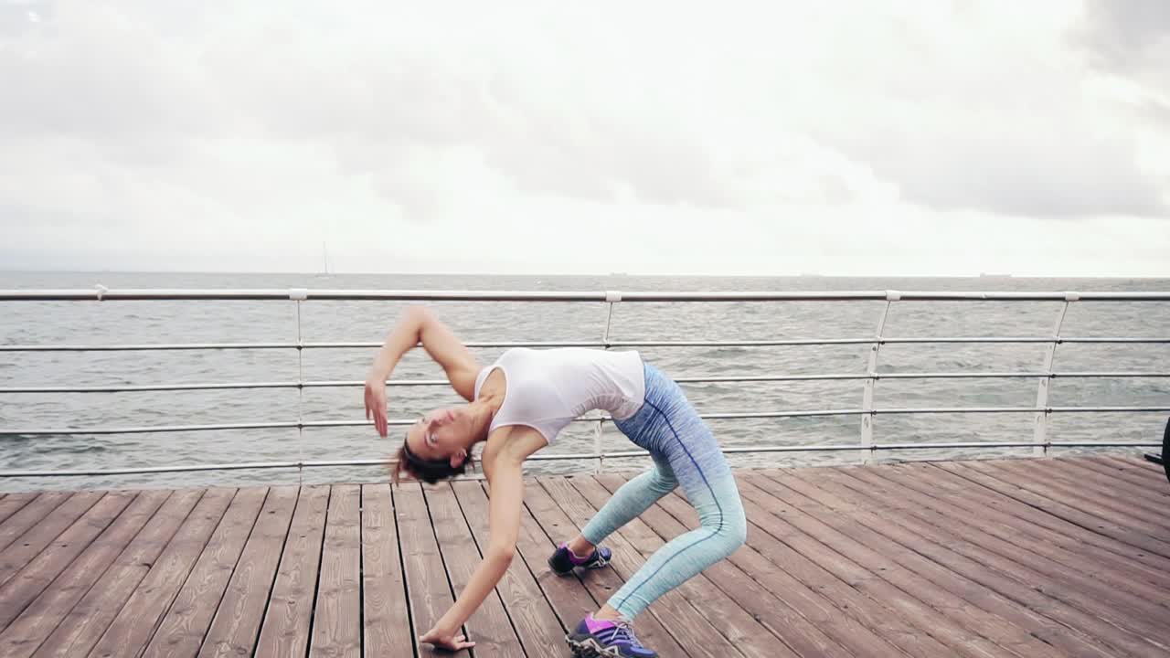 육상적인 젊은 여성이 바다에 의해 해변에 backbend 및 스트레칭을하고 있습니다. 느린 운동  ⁇