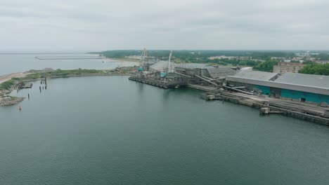 vista aérea de las grúas portuarias y los muelles de carga vacíos en el puerto de liepaja, la ciudad de liepaja en el fondo, día de verano nublado, amplia toma de avión no tripulado avanzando, inclinándose hacia abajo