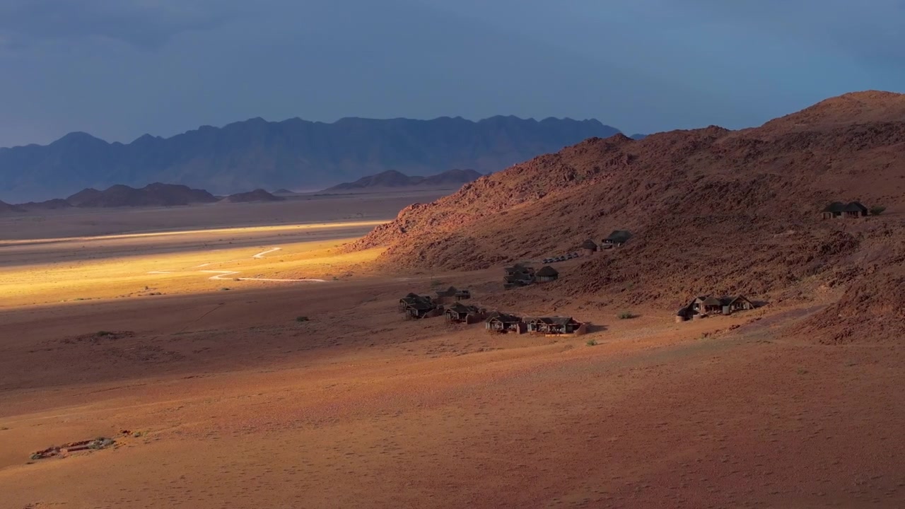 Drohnenansicht einer goldenen Dämmerung, die hinter den silhouettierten Bergketten der Namib-Wüste leuchtet. Sanftes Licht und treibender Nebel hüllen die alten Dünen und Berge in eine ruhige, filmische Atmosphäre.