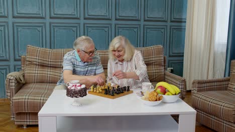 Retired-senior-couple-talking-drinking-tea,-playing-chess-in-modern-living-home-room-lounge-together