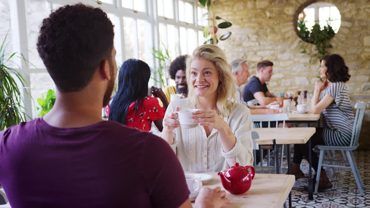 Young blonde woman holding coffee and talking to her boyfriend at a table by a window in a busy restaurant during the day, his back to camera