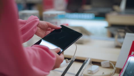 Lady seen from behind picking up a tablet from its stand in a store, tablet is connected with a security cord, while other tablets are arranged neatly on the table, she inspects the device closely