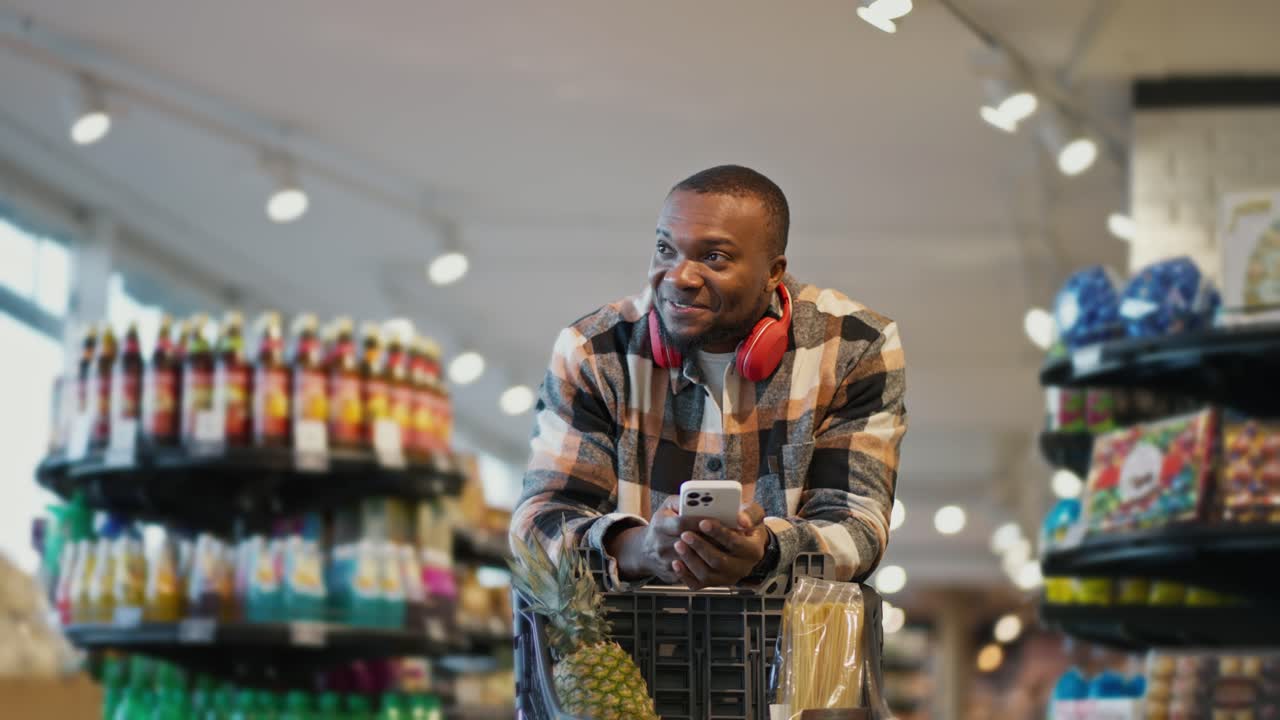 A happy man with Black skin and a short haircut and a beard in a checkered shirt looks at products on the shelves in a modern supermarket during his grocery shopping trip with cart