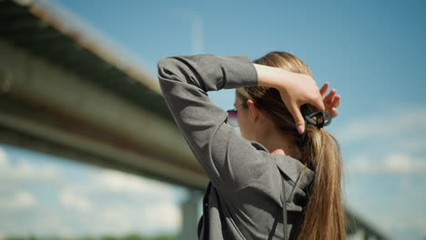 side view of lady wearing grey shirt as she uses a hair clip to gather her hair near a bridge, the background is slightly blurred, with the structure of the bridge