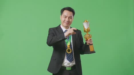 asian business man in a suit and tie with a gold medal and trophy touching his chest then smiling and pointing to camera being proud of himself on green screen background in the studio