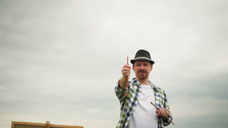 front view of an artist wearing a hat and a checked shirt, holding a painting brush and pointing it forward to determine an angle. the artist is focused, perfect composition in an outdoor setting