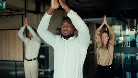 Ein-Schwarzer-Geschäftsmann-In-Einem-Weißen-Hemd-Macht-Yoga-Mit-Seinen-Kollegen-Im-Büro.-Machen-Sie-Eine-Pause-Von-Der-Arbeit,-Um-Spirituelle-Stärke-Und-Stabilität-Zu-Bewahren