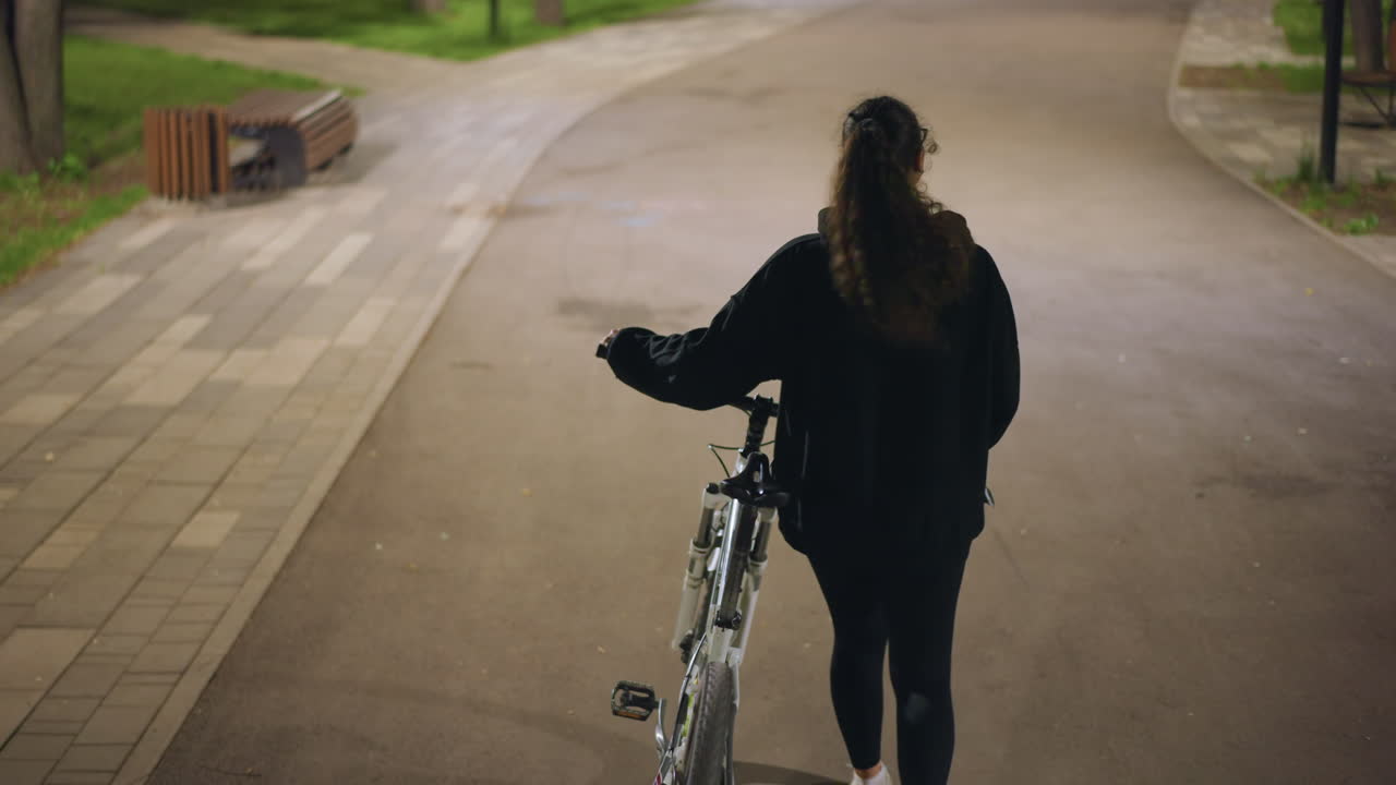 Woman Pedals Quiet Park, Caucasian Lady Cycling Along Tranquil Paved Pathway At Night, Woman With Ponytail Rides Her Bicycle Peacefully Through Softly Lit Park Pathway During Nighttime