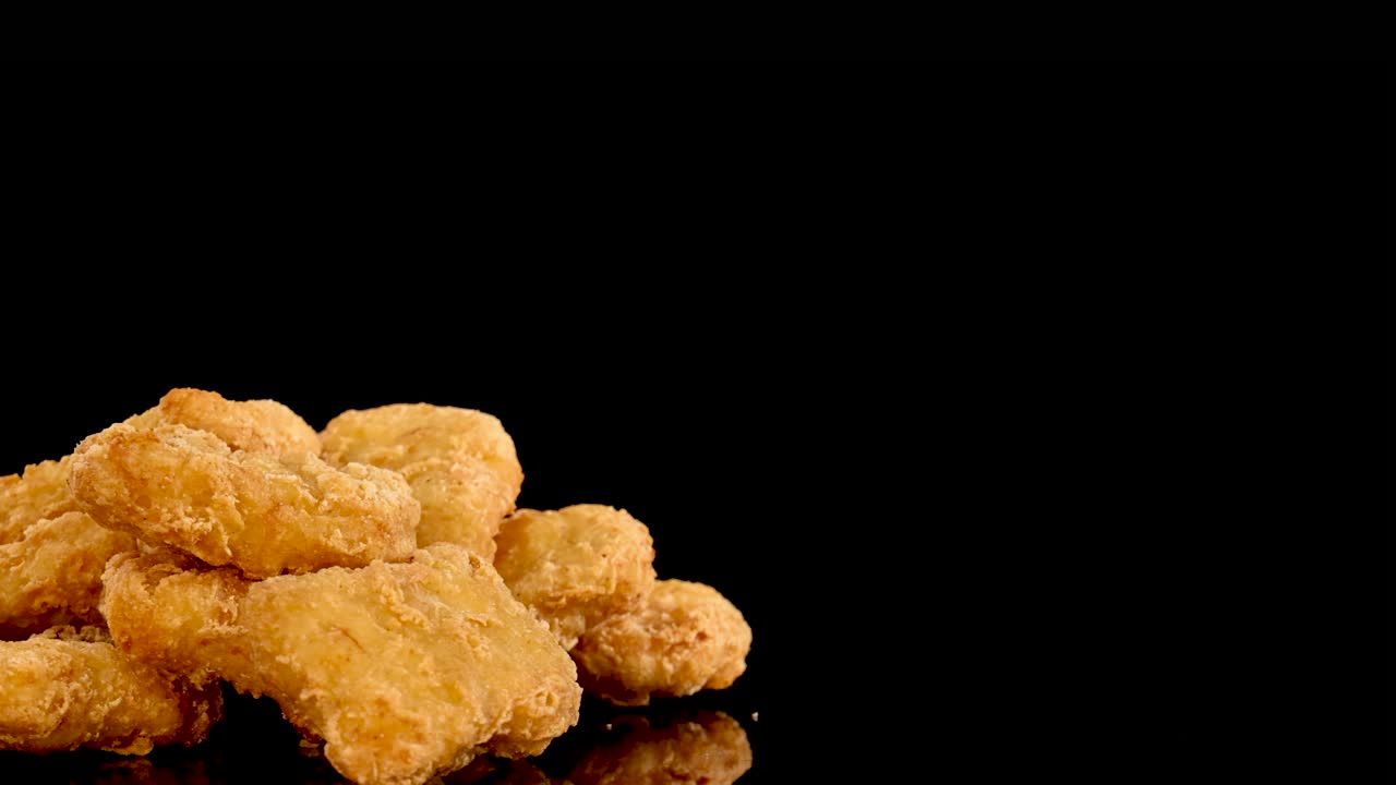 Golden, crumbed chicken nuggets rotate slowly on a reflective black surface under bright studio lighting, highlighting texture and appetizing appeal with dramatic close-up shots