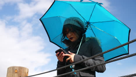 Mujer-Joven-Revisando-El-Clima-O-Enviando-Mensajes-De-Texto-En-Un-Teléfono-Inteligente-Con-Paraguas-Bajo-Un-Cielo-Azul-Con-Nubes-De-Lluvia-A-Cámara-Lenta