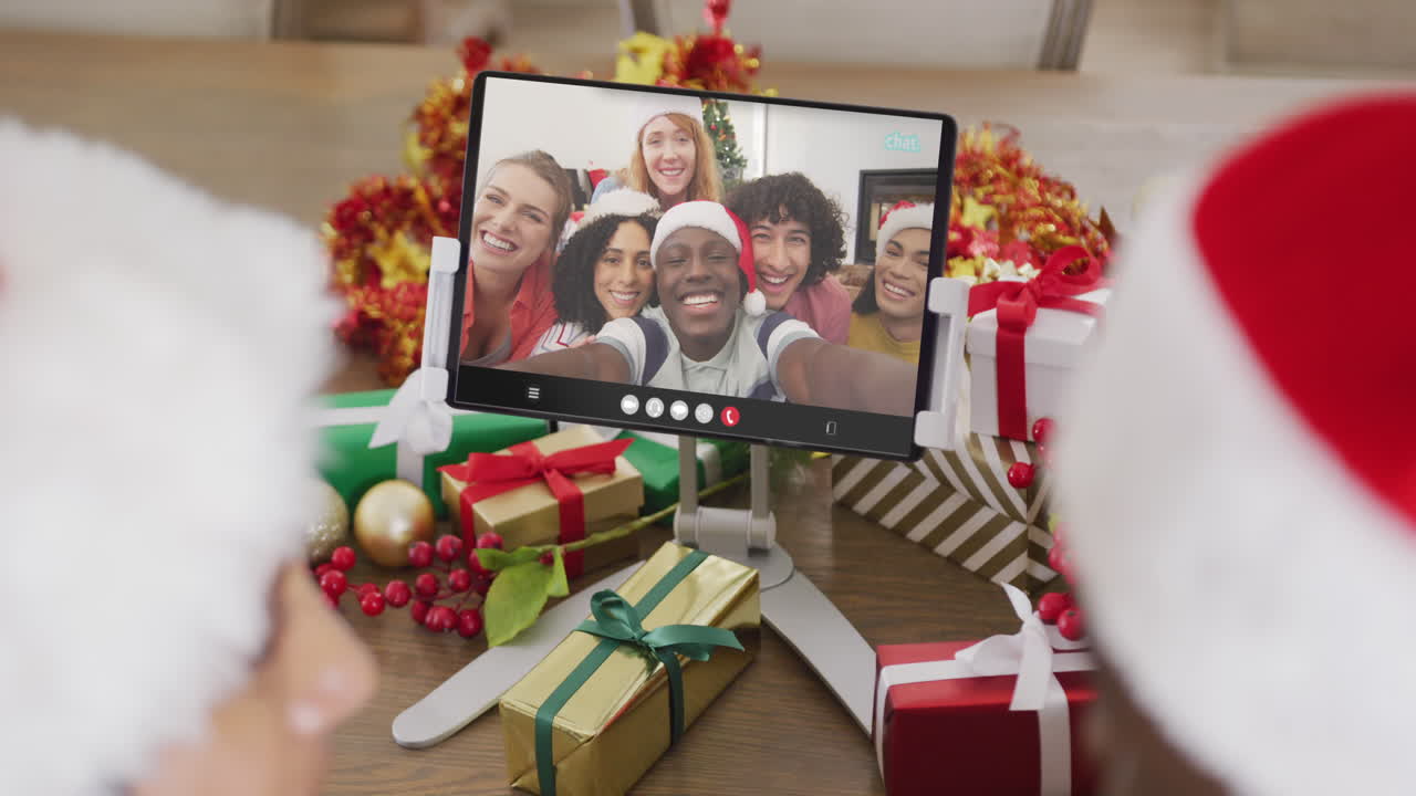 una pareja caucásica con sombreros de santa haciendo una llamada de video de tableta con amigos felices y diversos