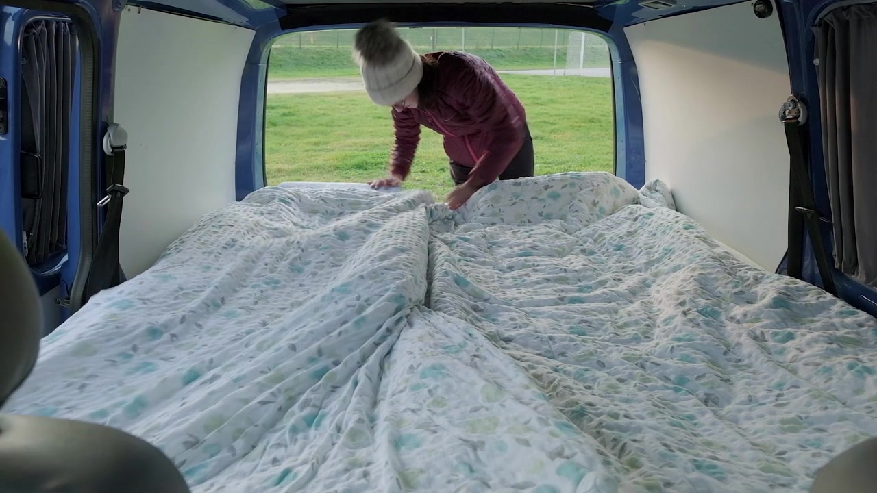A man and women making a bed in the back of a campervan. The camera set up looking into the back of the van with the back doors open. A couple travelling in a campervan.