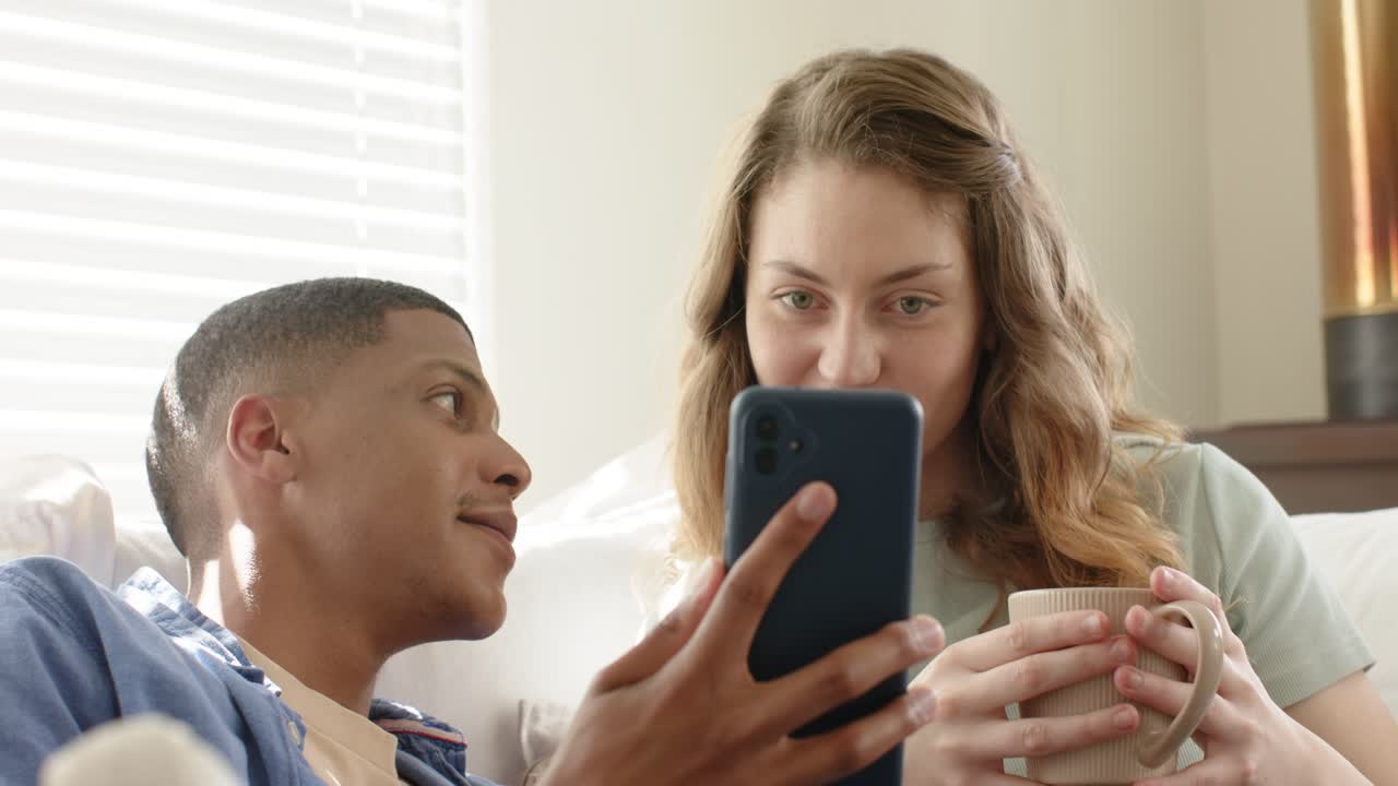 Casal diverso no sofá com caneca, girando o smartphone em direção a ela, examinando a tela e depois rindo