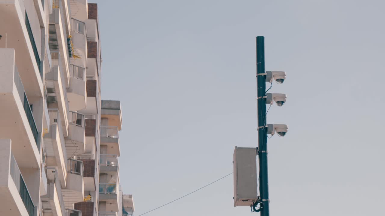 Tre telecamere di sorveglianza montate su un palo, con un edificio residenziale sullo sfondo, che simboleggiano gli sforzi della comunità contro l'insicurezza in un contesto urbano