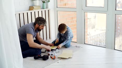 cute attentive boy is doing woodwork working with hammer and pliers while his experienced father is teaching him and measuring piece of wood with reel.