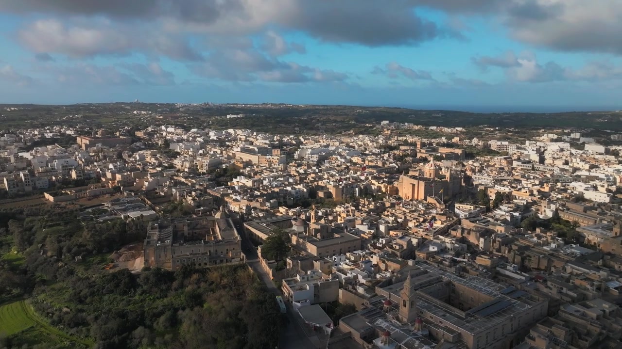 Una vasta ripresa aerea cinematografica della storica città fortificata di Mdina e Rabat a Malta durante l'ora d'oro, caratterizzata da architettura in pietra calcarea e campi verdi ondulati.