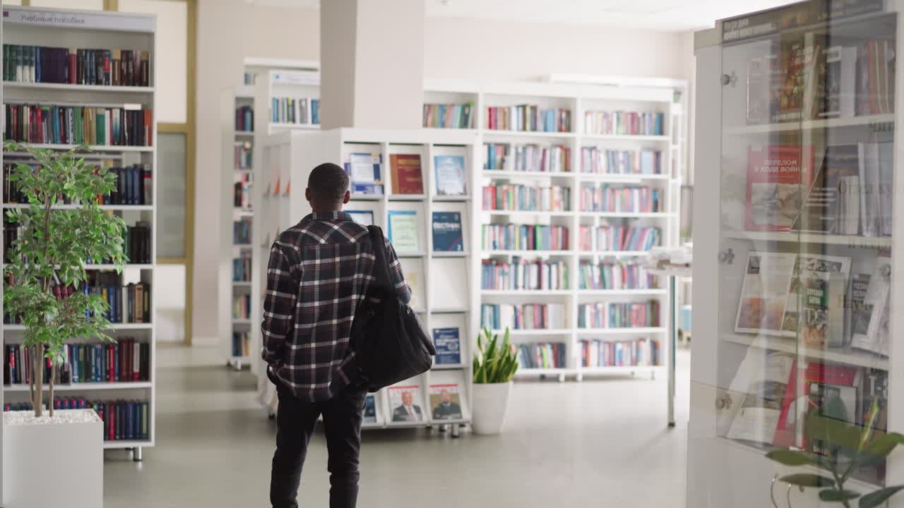 Man walks across public library hall. African American visitor with backpack comes for scientific books to literature storage. Reader at bookstore