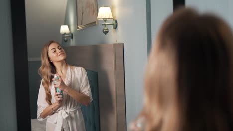 beautiful long-haired blonde woman sprays her hair in front of a mirror