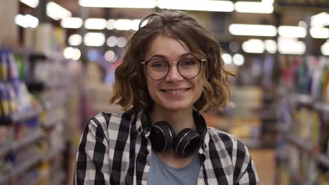 Portrait-young-woman-stands-in-front-of-the-camera-and-smiles-in-supermarket-feel-happy-girl-shopping-face-retail-store.-Pretty-curly-female-customer-in-casual-with-headphones-on-neck