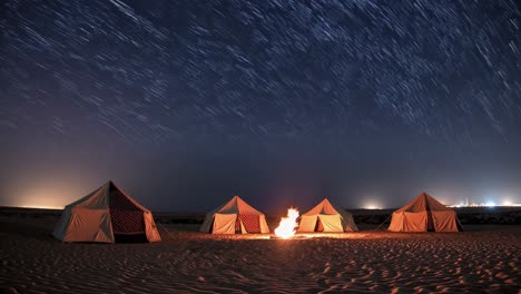 four tents are set up in the desert sand, illuminated by a warm campfire in the foreground, under a mesmerizing night sky filled with star trails, creating a tranquil and adventurous camping scene
