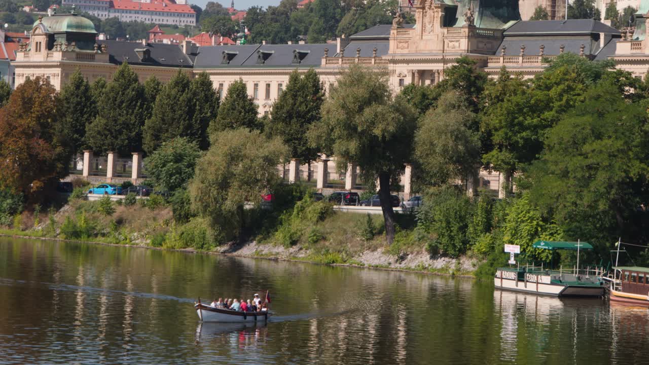 Piccole file di gruppo su un fiume calmo con paesaggio urbano storico, alberi rigogliosi e luce diurna intensa